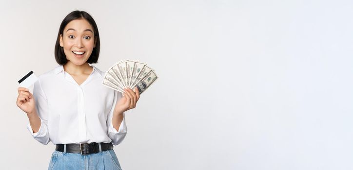 Portrait of enthusiastic asian woman holding money in cash and credit card, smiling amazed at camera, white background.