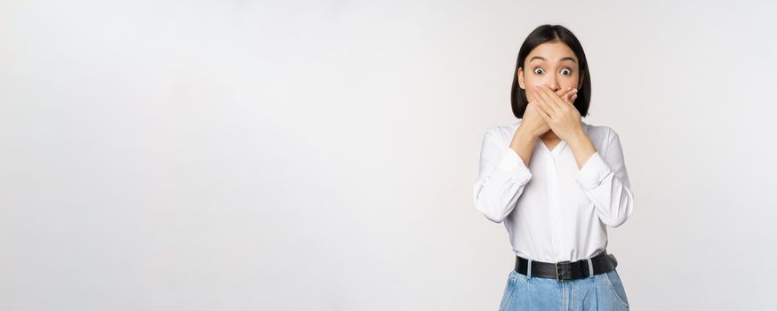 Shocked young asian girl close mouth, cover lips with hands and looking amazed, standing over white background. Taboo gesture