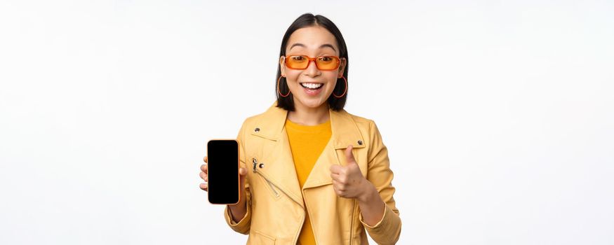 Beautiful korean girl, asian woman in sunglasses, showing smartphone app interface, thumbs up, recommending mobile phone application, white background.