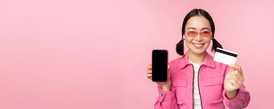 Image of smiling korean woman showing credit card and mobile phone screen, smartphone application interface, paying online, shopping contactless, standing over pink background.