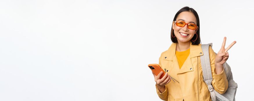 Young asian woman traveller, girl tourist in sunglasses, holding backpack and mobile phone, using application on smartphone, standing over white background.