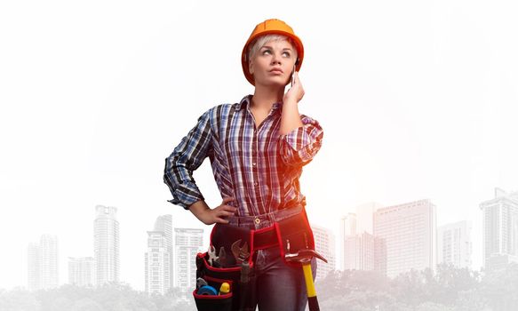 Attractive blonde woman in workwear and hardhat talking on smartphone. Portrait of young worker in checkered blue shirt standing on foggy downtown background. Business communication and conversation.