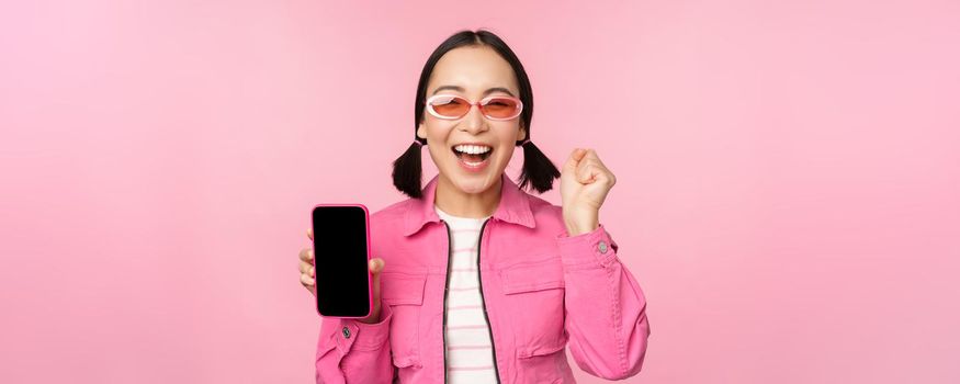 Excited asian girl laughs and smiles, shows mobile phone screen, smartphone application, standing over pink background.