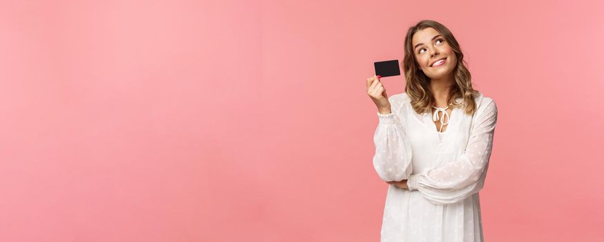 Portrait of cute tender feminine blond girl daydreaming about how she will waste her money on online shopping, look up dreamy, imaging as waiting delivery, hold credit card, pink background.