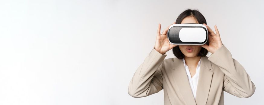 Amazed office woman, asian business person in suit, wearing vr headset, looking at smth in virtual reality glasses with impressed, wow face, white background.