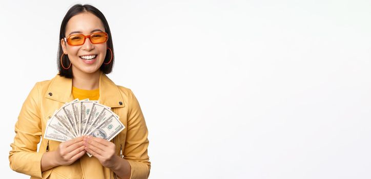 Stylish smiling asian girl holding money cash, showing dollars and celebrating, standing over white background. Copy space