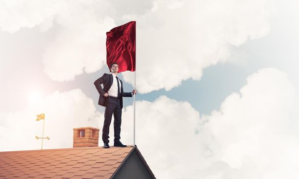 Businessman standing on house roof and holding red flag. Mixed media