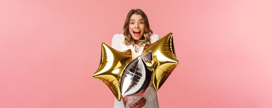 Holidays, celebration and women concept. Happy charming and surprised girl being congratulated with birthday, holding star-shaped balloons and smiling joyfully camera, pink background.