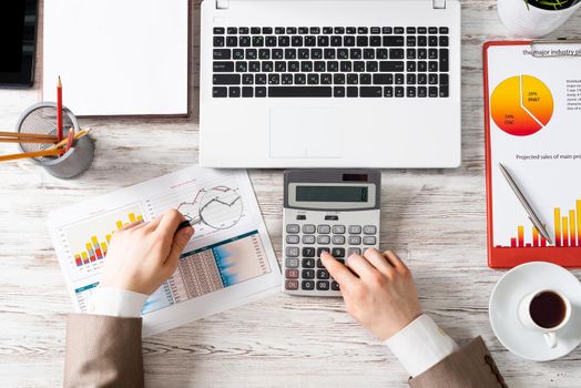 Businessman working at office desk with financial data. Top view office workplace with human hands using calculator. Finance accounting concept. Flat lay table with laptop and business charts.