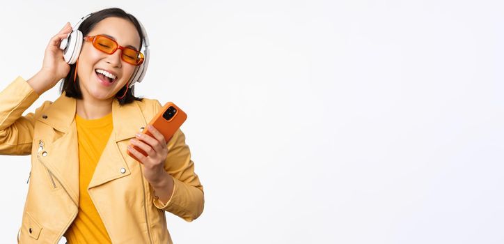 Technology concept. Stylish asian girl in headphones, holding smartphone, dancing and singing, listening music, standing over white background.