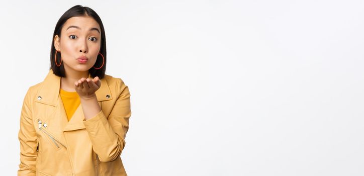 Cute korean stylish girl, sending air kiss, blowing mwah and looking silly romantic at camera, standing over white background. Copy space