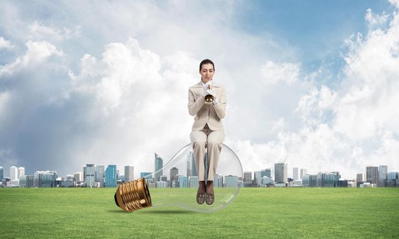 Woman sitting on big light bulb and playing trumpet brass. Young lady in white business suit and gloves with music instrument on green field. Business assistance and support concept with musician.