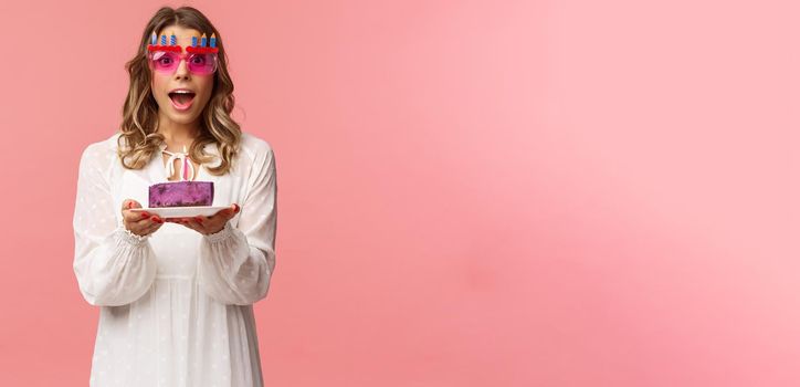 Holidays, spring and party concept. Excited, happy attractive blond woman in white dress, wearing funny b-day glasses, holding birthday cake with lit candle, making wish smiling camera.