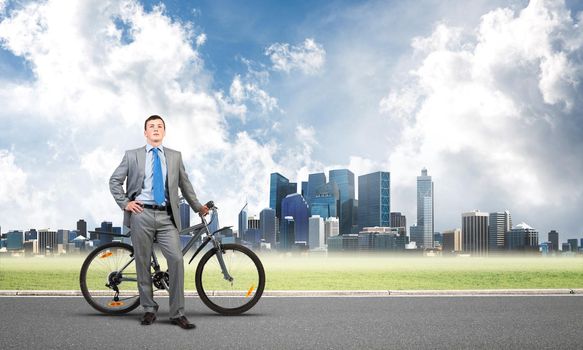 Young man wearing business suit and tie standing on asphalt road with bike. Businessman with bicycle on background of business center. Outdoors male cyclist holding bicycle, having break in riding.