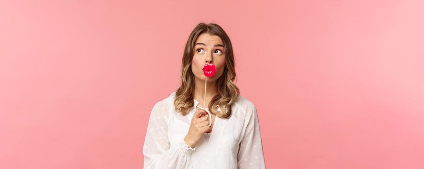 Spring, happiness and celebration concept. Close-up of lovely, dreamy and romantic, tender blond girl in white dress, holding lips on stick and looking up thoughtful, wondering over pink background.