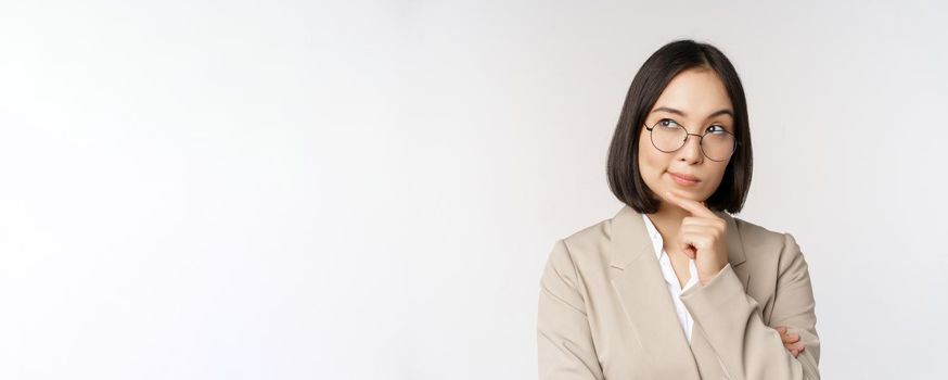 Portrait of thoughtful asian businesswoman in glasses, making assumption, thinking, standing in beige suit against white background.