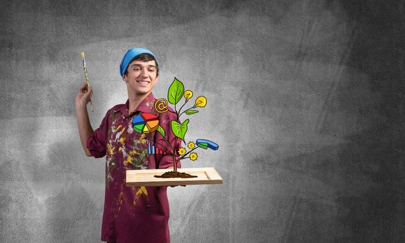 Young artist drawing plant of wealth and business success. Happy painter in dirty shirt and bandana standing on grey wall background. Original business idea generation and development.