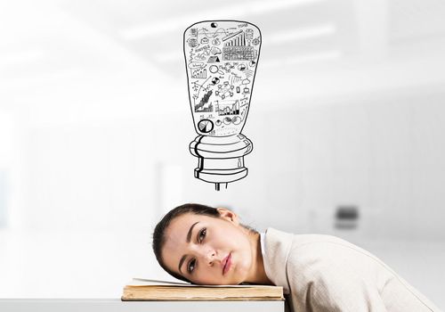 Bored business woman lying on desk with open book. Young tired student trainee in white business suit relaxing in light office interior. Idea light bulb sketch with business doodles above head.