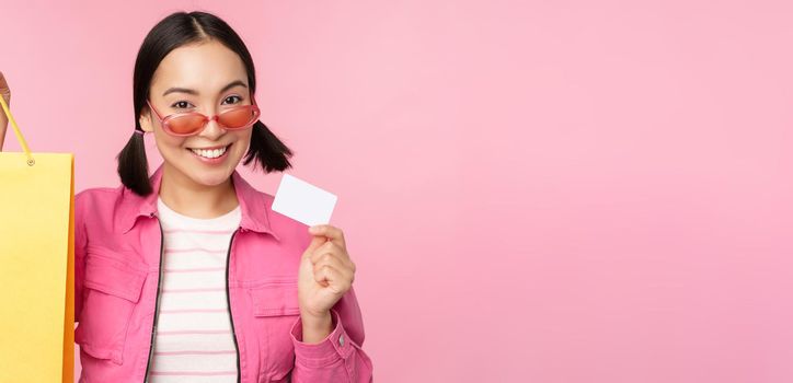 Happy young asian woman shows credit card and shopping bag, store sale announcement, buying smth in shop, posing against pink background.