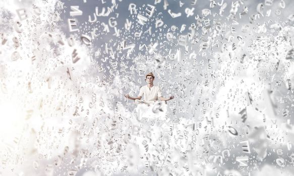 Man in white clothing keeping eyes closed and looking concentrated while meditating among flying letters in the air with cloudy skyscape on background.
