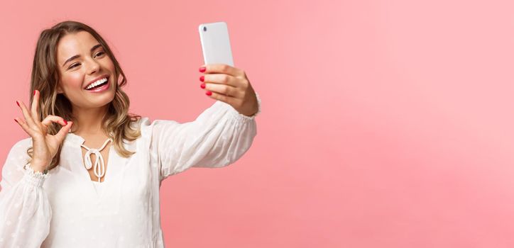 Close-up of satisfied good-looking blond girl in white dress, taking selfie, record mobile phone video, show okay satisfactory sign with pleased nod, smiling agree or recommend, pink background.