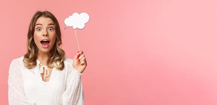Spring, happiness and celebration concept. Close-up portrait of surprised, excited blond girl looking amazed, holding cloud comment stick near head and gasping astonished, have idea, pink background.