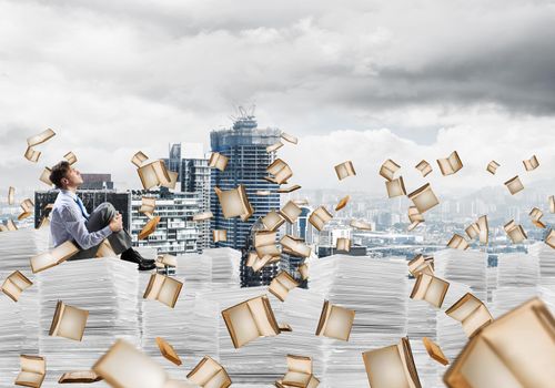 Businessman looking away while sitting on pile of documents among flying books with cityscape on background. Mixed media.