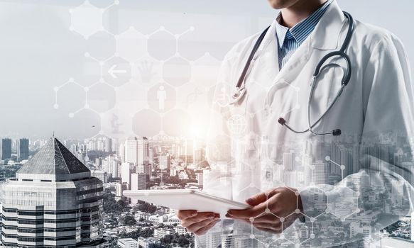 Confident female doctor in white sterile coat using tablet while standing outdoors with cityscape on background. Medical industry concept. Double exposure with interface icons