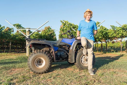 Young farmer standing near quad bike on background of vine rows. Traditional farming in vineyard. Blond guy in straw hat and and blue t-shirt outdoor. Ecological food production in countryside.
