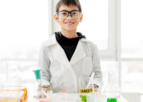 School boy wearing protection glasses doing chemistry experiment in elementary science class. Clever pupil looking at camera and smiling in lab during test