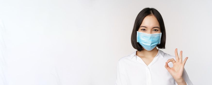 Covid and health concept. Portrait of asian woman wearing medical face mask and showing okay sign, standing over white background.