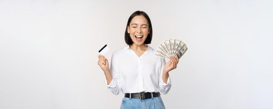 Finance and money concept. Happy young asian woman dancing with cash and credit card, smiling pleased, posing against white studio background.