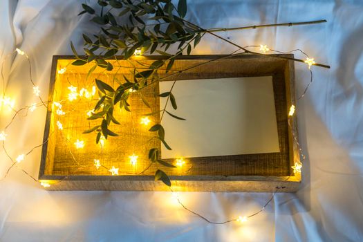 A eucalyptus branch lies on a white sheet beside a tray with a letter in an envelope. The garland is glowing. Copy space. Place for text.