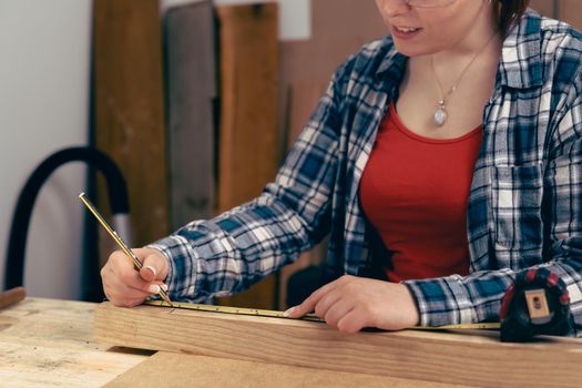 Young carpenter with red hair, measuring a wooden board with a tape measure in her small carpentry workshop, dressed in blue checked shirt and red t-shirt. Young businesswoman working by hand on a piece of wood and designing new furniture for the house. Warm light indoors, background with wooden slats. Horizntal.