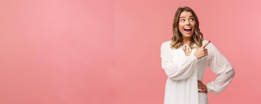 Enthusiastic, happy attractive blond woman in white cute dress, open mouth excited and cheerful looking, pointing upper right corner as best prices discounts ever, stand pink background.