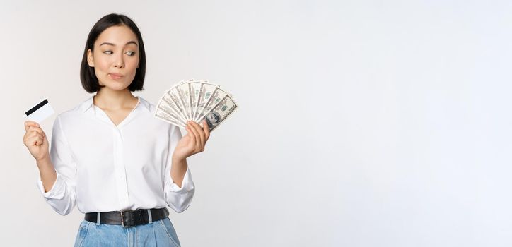 Image of asian woman looking at money dollars, holding credit card in another hand, thinking, standing over white background.
