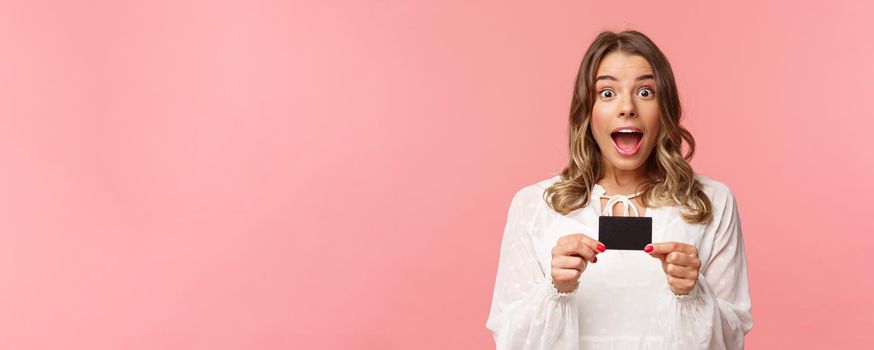 Close-up portrait of emotive, excited and thrilled good-looking blond girl showing credit card and feeling very happy, telling about discounts, special offers, bank service, pink background.