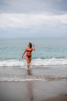 A middle-aged woman with a good figure in a red swimsuit on a pebble beach, running along the shore in the foam of the waves.
