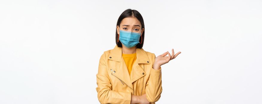 Confused asian woman in medical face mask, looking clueless, puzzled, wearing medical face mask, standing over white studio background.