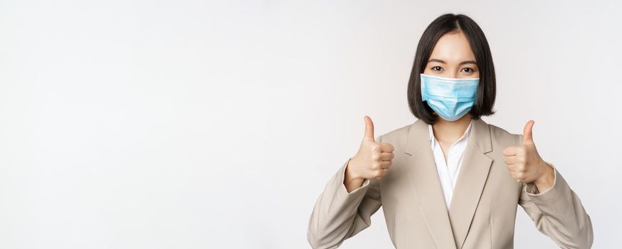 Coronavirus and workplace concept. Image of asian saleswoman, company worker in medical mask showing thumbs up sign, smiling pleased, white background.