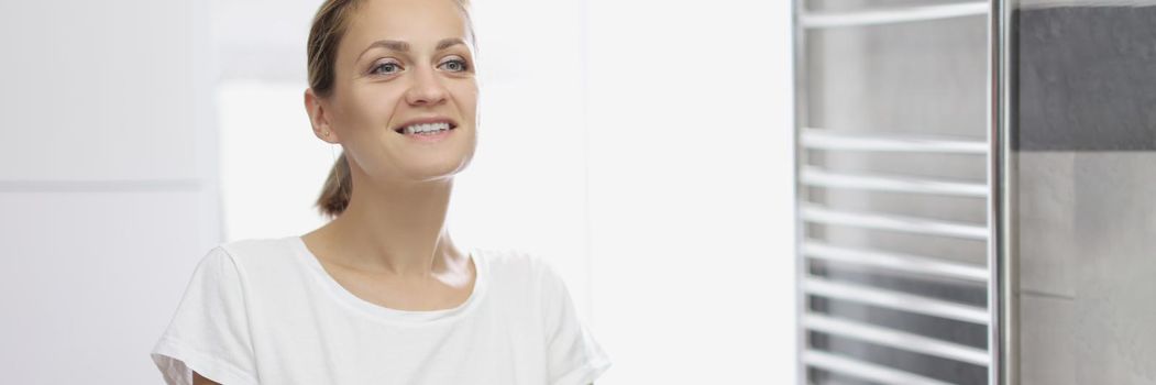 Portrait of young woman looking in mirror at herself in bathroom, white smile, natural beauty, no makeup, morning routine, light restroom. Good morning, wellness, beauty, youth concept