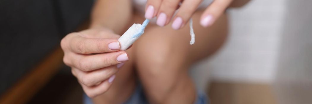 Close-up of woman hold tampon, feeling sick during critical days, hygiene care. Girl sit on toilet in bathroom. Menstrual cycle, gynecological health, protection, hygiene concept. Blurred background