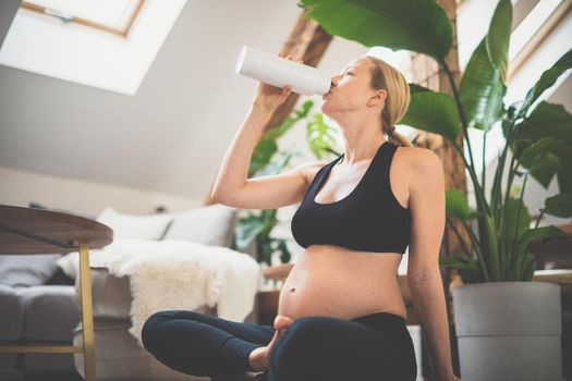 Young happy and cheerful beautiful pregnant woman taking a break, hydrating, drinking water from the botle after home well being workout program