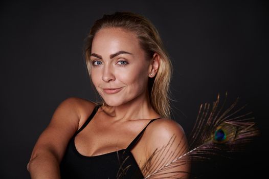 Close-up gorgeous smiling stunning middle aged blonde Caucasian woman with healthy fresh clean skin and wet blond hair holding a peacock feather, isolated over black background with copy ad space