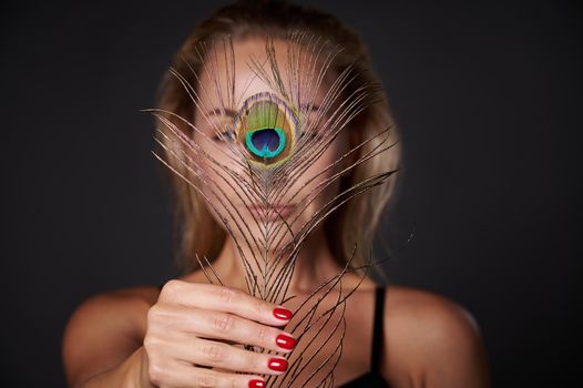 Focus on the peacock feather in the hands of a beautiful blonde woman, isolated over black background with copy space for ads
