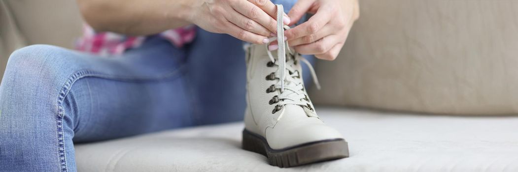 Close-up of woman sit on couch and tie shoelaces on boots, female placed foot on clean sofa. Fix shoes before going out. New footwear, purchase, shopping, fashion, style concept