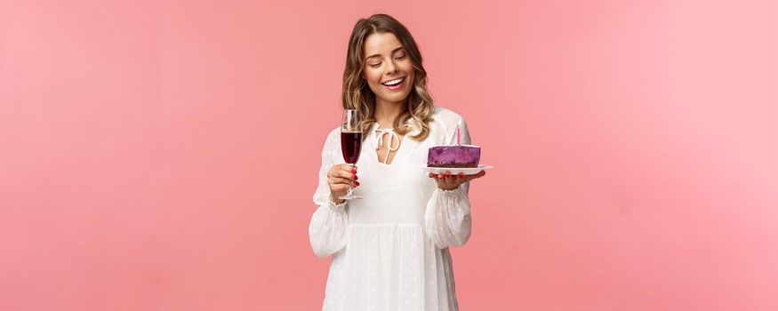 Holidays, spring and party concept. Portrait of tender, elegant young blond woman holding glass of wine and cake with lit candle, smiling pleased, celebrating birthday, pink background.