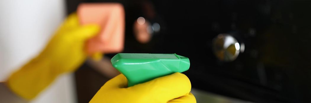 Close-up of woman housekeeper wiping dust from oven using spray and fabric, keep surface clean and shiny at home. Cleaning service job, cleanup, sanitizing, clean space concept. Blurred background