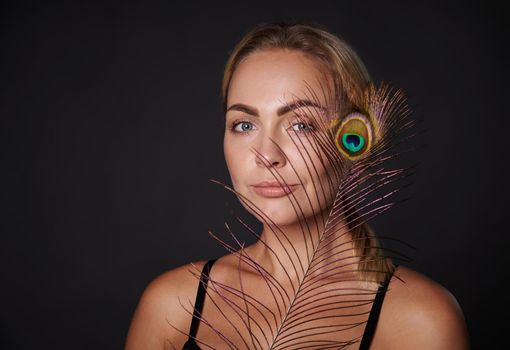 Beautiful middle aged blonde woman with healthy fresh clean skin holding a peacock feather, isolated over black background with copy ad space