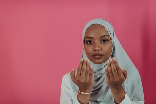 Modern African Muslim woman makes traditional prayer to God, keeps hands in praying gesture, wears traditional white clothes, has serious facial expression, isolated over plastic pink background. High-quality photo